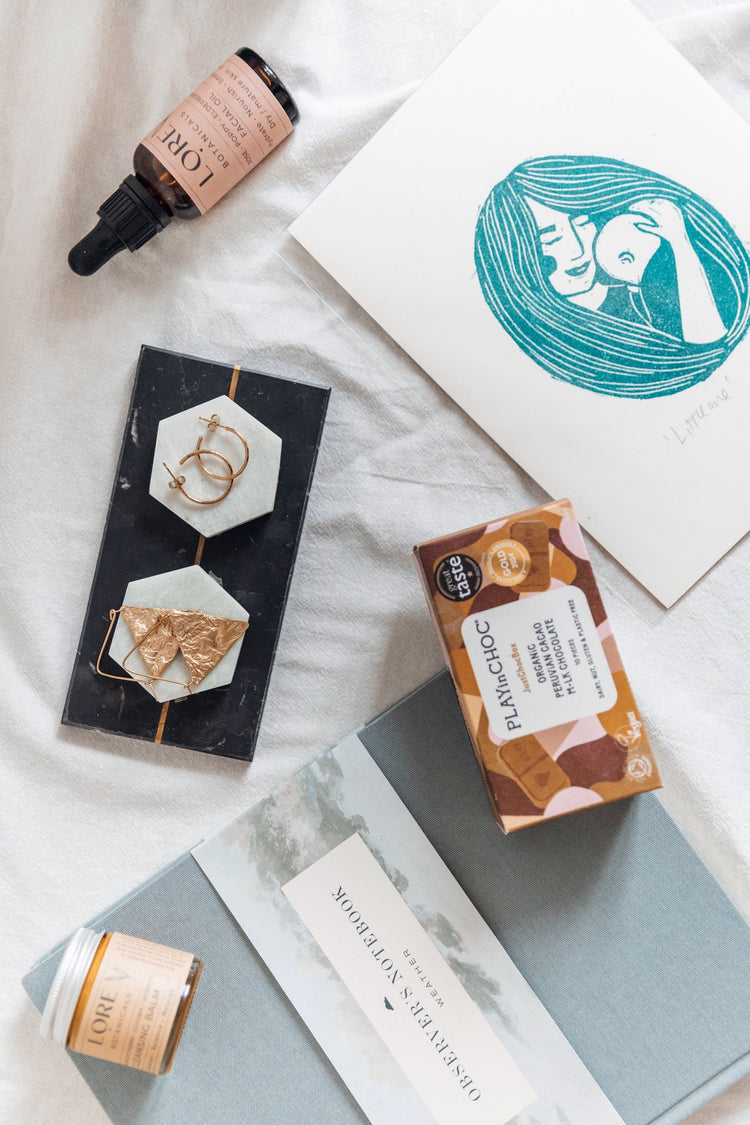 flat lay of a print, face oil, chocolates, jewellery  and a notebook