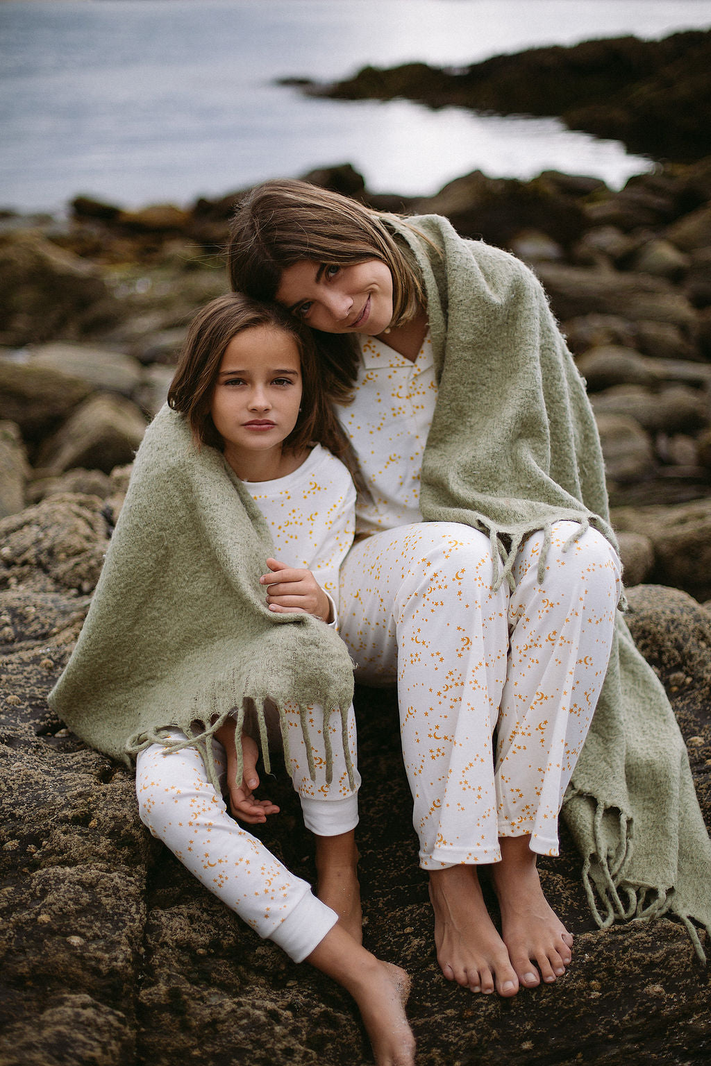 a girl and a woman sat on a rock at the sea side wearing organic sleepy doe pyjamas and wrapped in a blanket