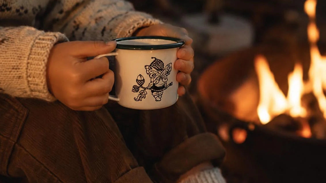 A cozy lifestyle shot of a person sitting by a campfire, holding a white enamel mug with a black floral design. The person is wearing a cream knit sweater, brown pants, chunky knit socks, and brown shoes.